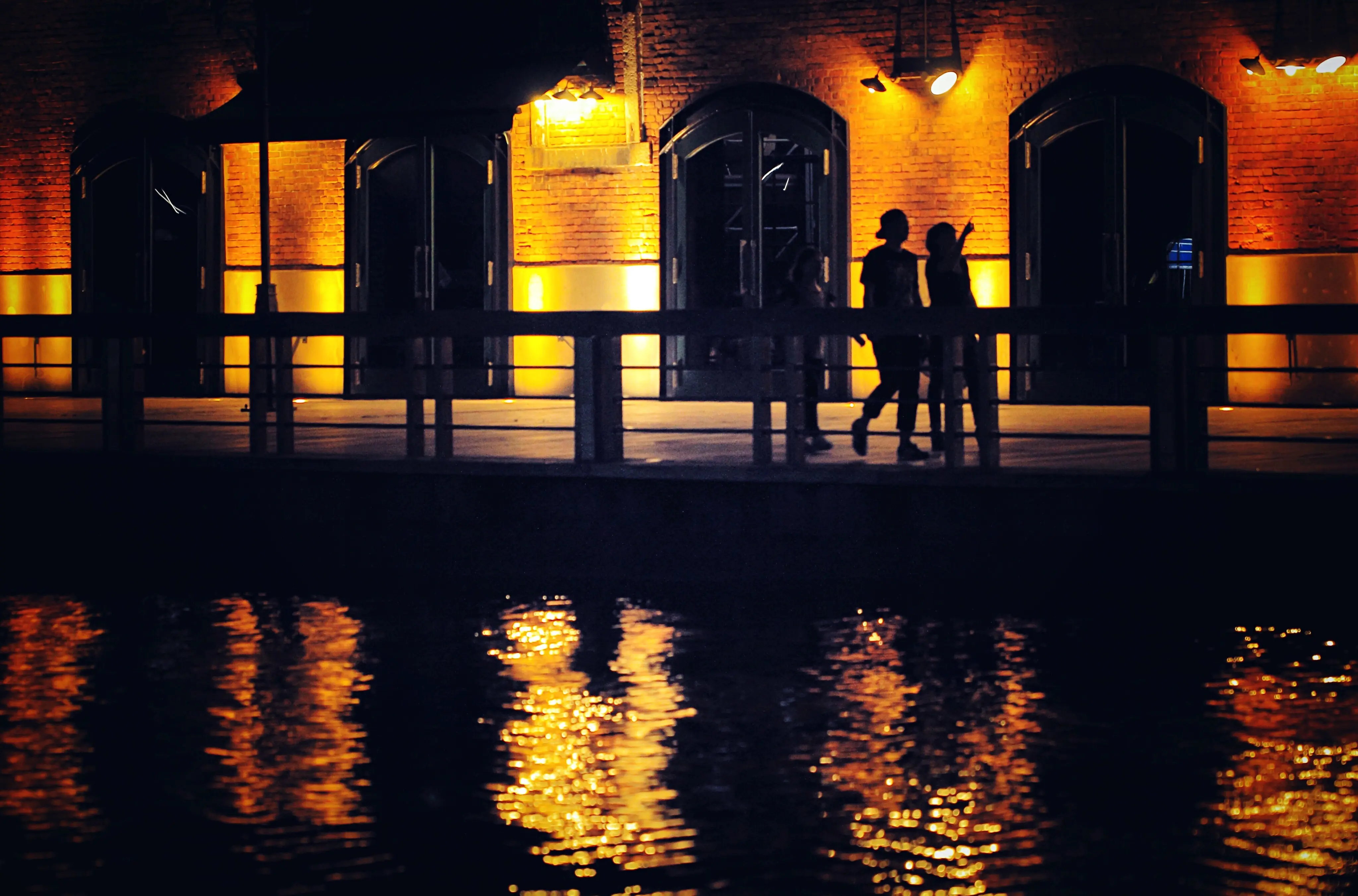 Silhouetten von Menschen, die am Fluss entlanggehen, vor einem beleuchteten Gebäude bei Nacht.